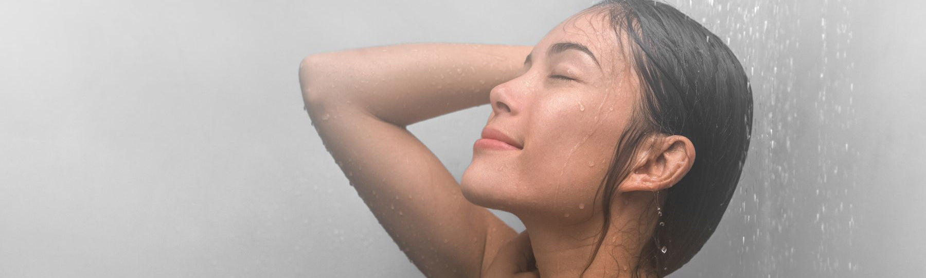 A woman with wet hair stands under a shower with her eyes closed and hand behind her head, enjoying the water streaming down her face and shoulders. The background is misty and grey.