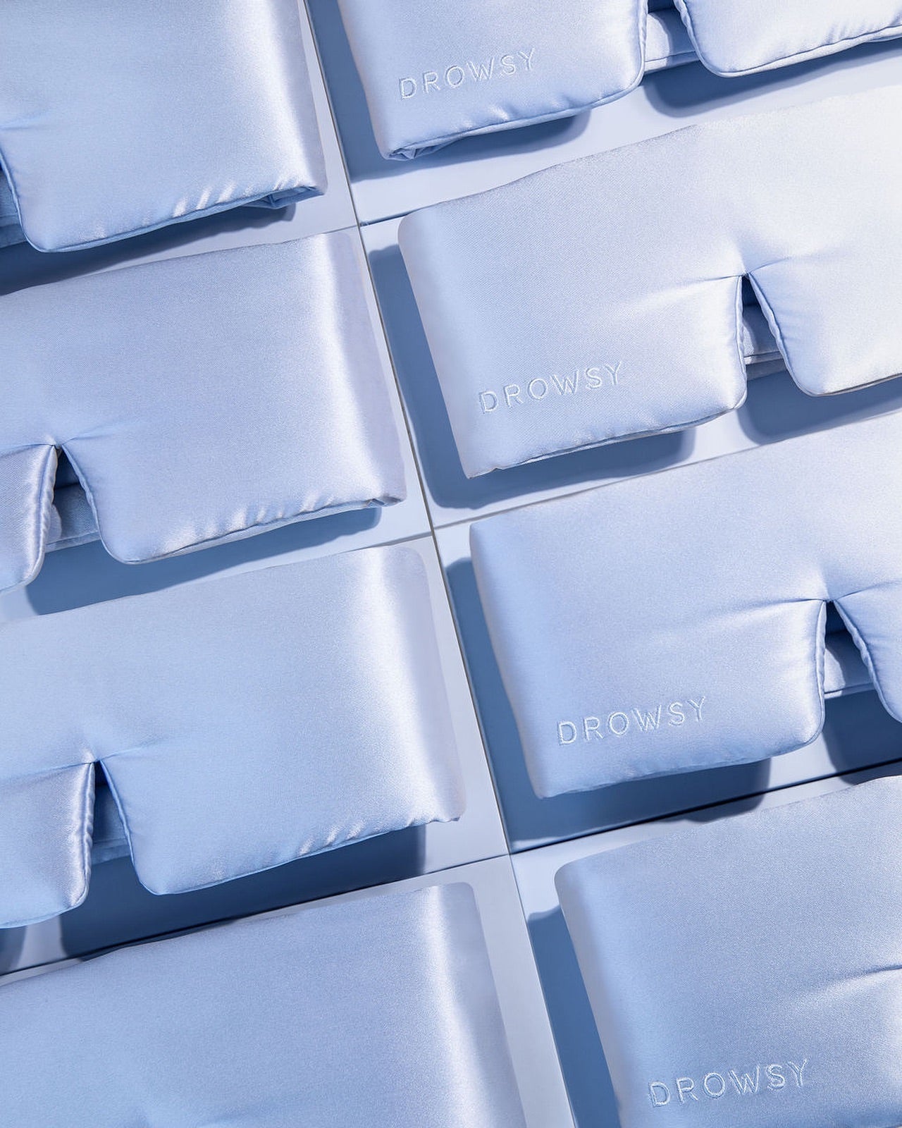 A flat lay of multiple pale blue satin sleep masks, each embroidered with the word DROWSY, arranged neatly in rows on a light surface.