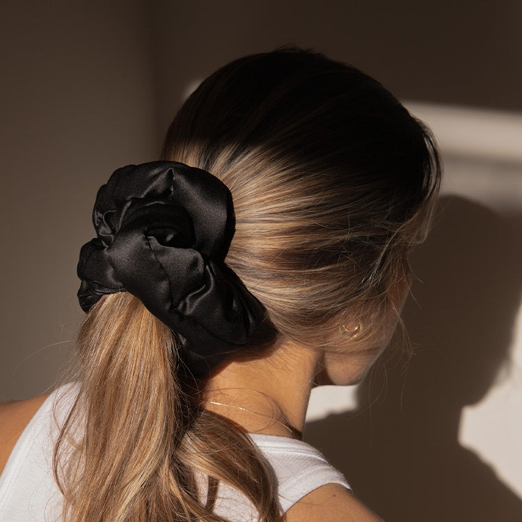 A woman with blonde hair tied in a low ponytail, secured with a large black satin scrunchie, is seen from behind. She is wearing a white sleeveless top and small gold earrings, standing in soft, natural light.