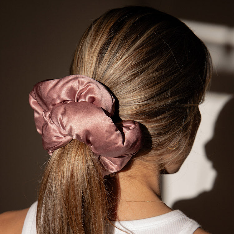 A woman with light brown hair pulled back in a low ponytail, secured with a large, shiny mauve satin scrunchie. She is wearing a white sleeveless top and is facing away from the camera.