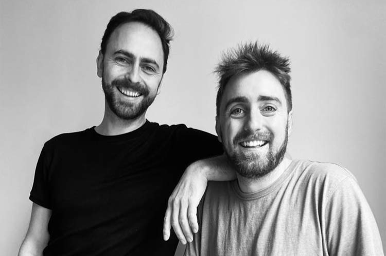 Two men smiling at the camera. One, wearing a black t-shirt, stands with his arm resting on the shoulder of the other, who is seated and wearing a light-colored shirt. Both have short hair and beards, set against a plain background.