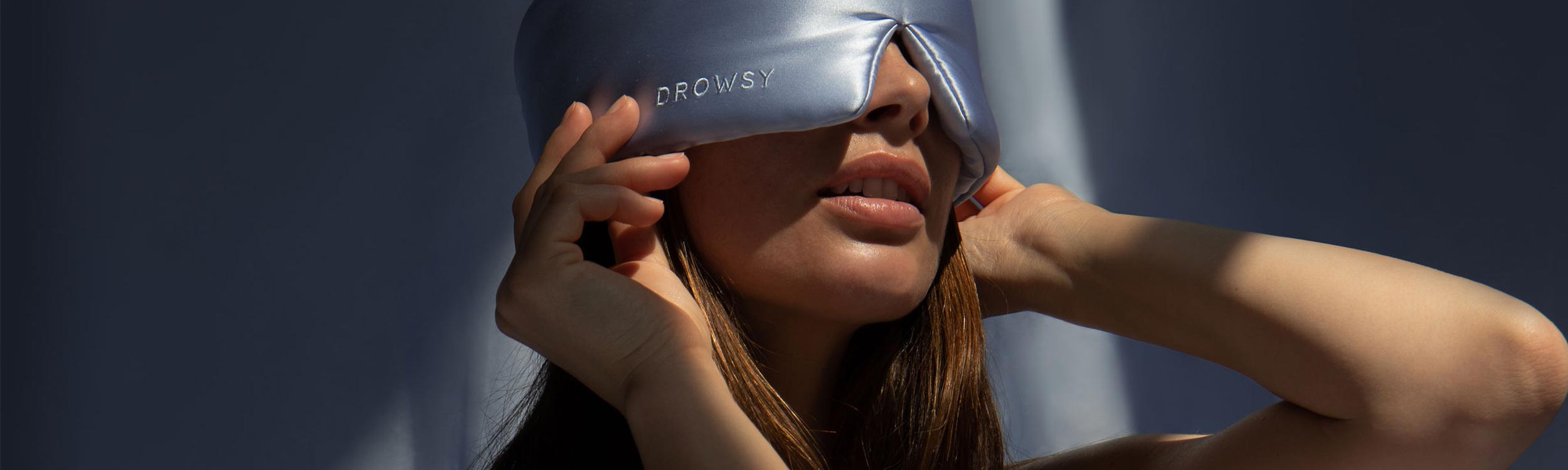 A woman adjusts a silky, light gray sleep mask labeled “Drowsy” over her eyes, with sunlight casting shadows across her face and background.