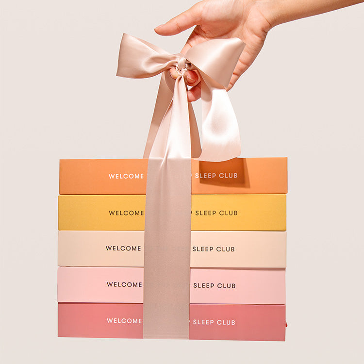 A hand holds a stack of five colorful rectangular boxes tied together with a large, pale pink satin ribbon. Each box is labeled WELCOME TO THE SLEEP CLUB in white text.