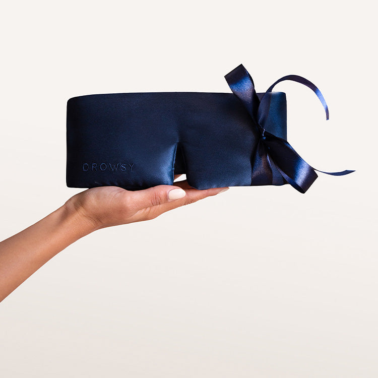 A hand with painted nails holds a dark blue silk sleep mask with a large ribbon bow, against a plain light background. The word DROWSY is subtly embossed on the mask.