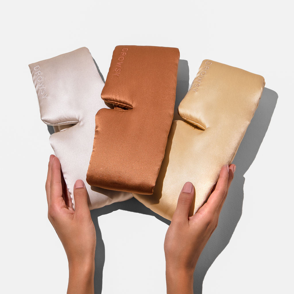 Two hands hold the Drowsy Besties Pack: three luxury mulberry silk sleep masks in beige, copper, and gold on a white background. Each mask is embroidered with “DROWSY,” offering the ultimate in beauty sleep.