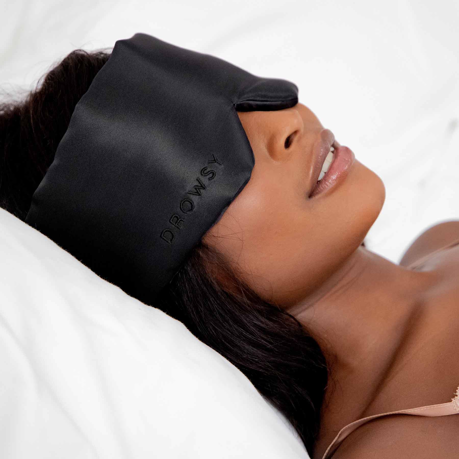 A woman lies on a white pillow wearing a black satin sleep mask with the word DROWSY embroidered on it, relaxing with a peaceful expression.