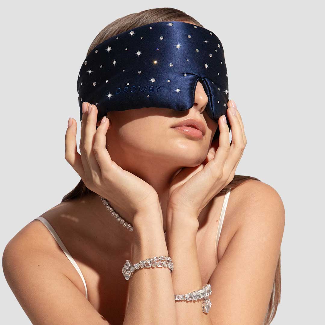 A woman in a white spaghetti strap top wears the us.drowsysleepco Silk Sleep Eye Mask Crystals by Swarovski®, featuring a navy 100% blackout star-patterned design, and accessorizes with Swarovski crystals on her neck, wrist, and fingers.