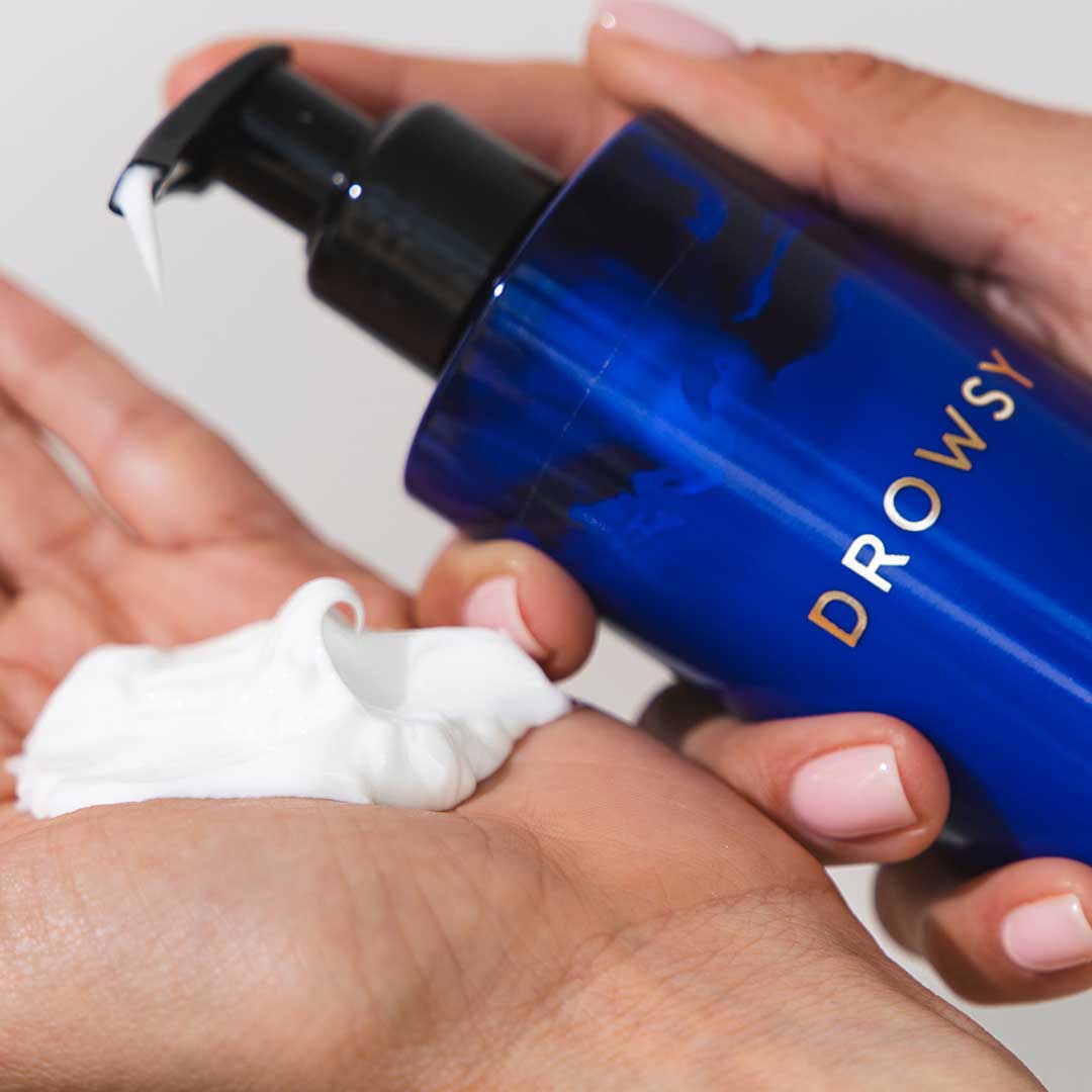 A person dispenses foamy white Beauty Sleep Body Cream from a blue us.drowsysleepco bottle onto their palm, revealing neatly manicured nails. This formula helps you relax and unwind before bed.