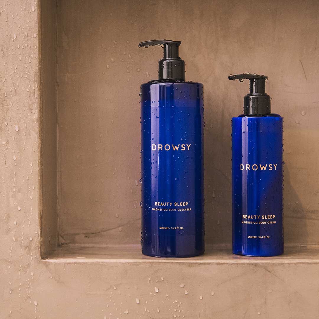 Two blue bottles of the us.drowsysleepco Beauty Sleep Ritual—a larger body cleanser and a smaller magnesium-infused body cream, both with pump dispensers—rest on a wet shower shelf. Water droplets dot the beige shower walls.