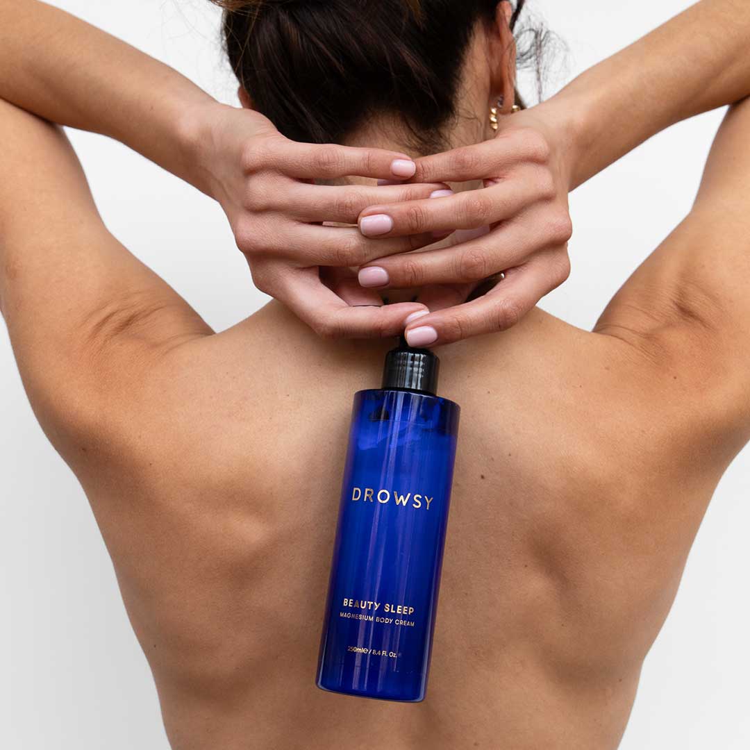 A person with their back to the camera holds their arms raised behind their head, displaying a blue bottle labeled Drowsy Beauty Sleep resting on their upper back.