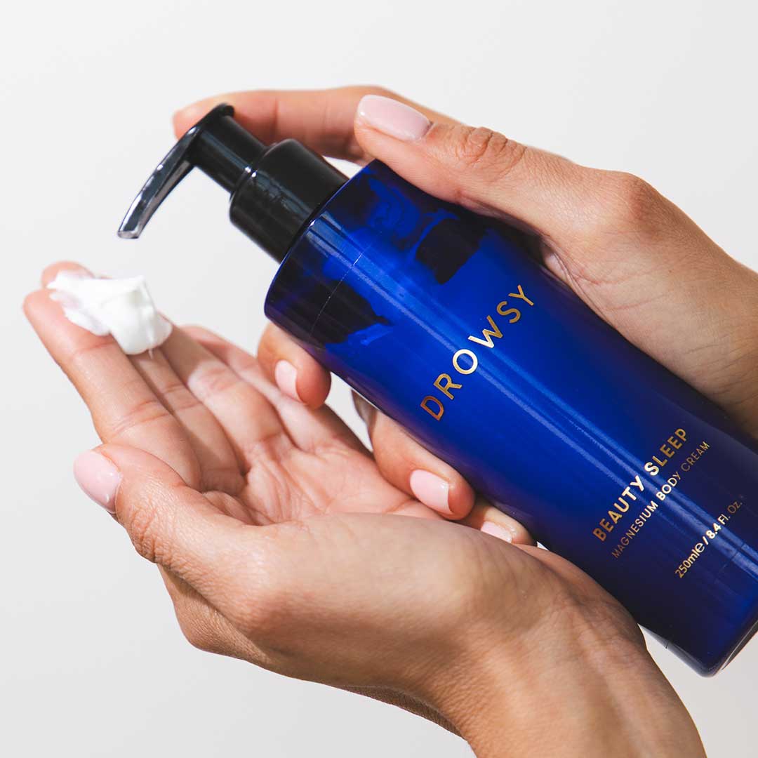 A person dispenses Beauty Sleep Ritual cream by us.drowsysleepco from a blue pump bottle onto their hand, showcasing this magnesium-infused body care as part of a nightly sleep ritual. The background is plain white.