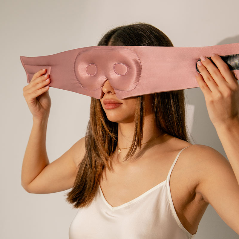 A woman in a white camisole puts on a large pink silk eye mask, preparing to cover her eyes. She stands against a plain light-colored background.