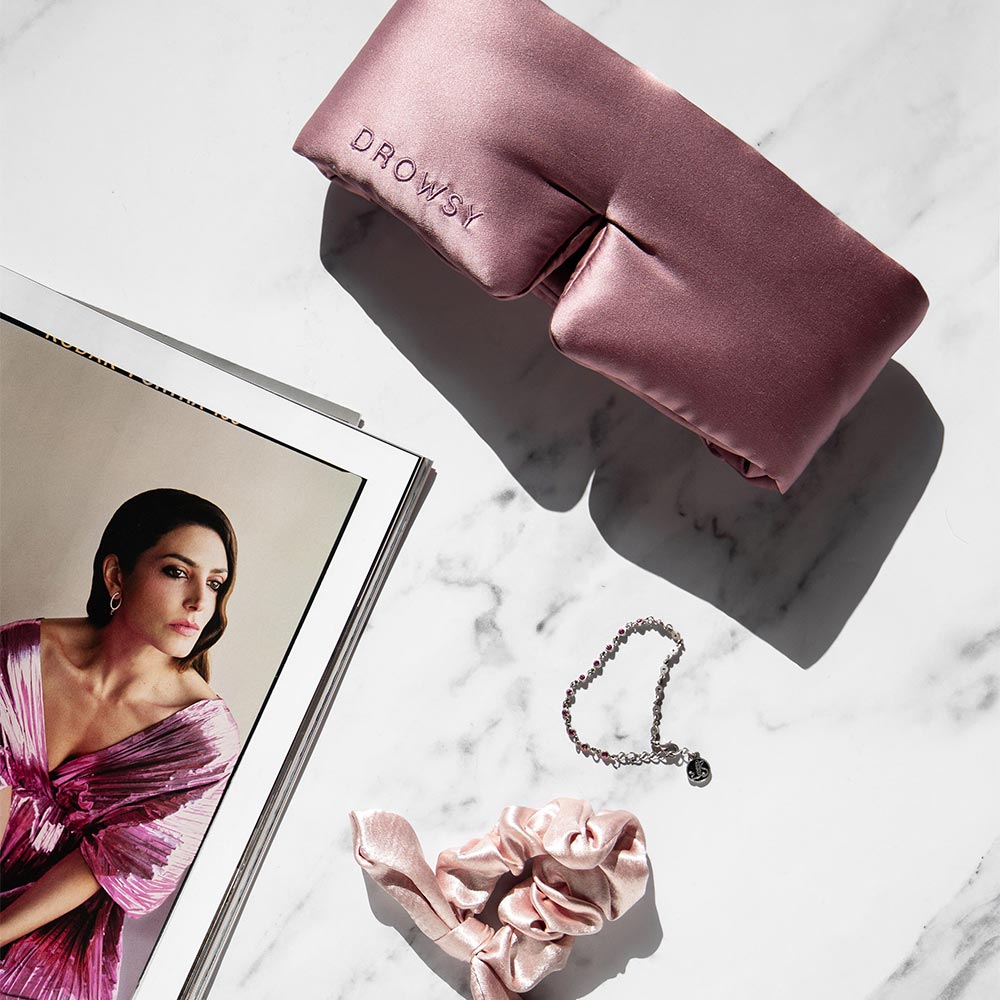 The Damask Rose set from us.drowsysleepco features a pink silk blackout eye mask, matching scrunchie, and silver bracelet, all arranged on white marble beside a magazine—perfect for relaxation and skin hydration.