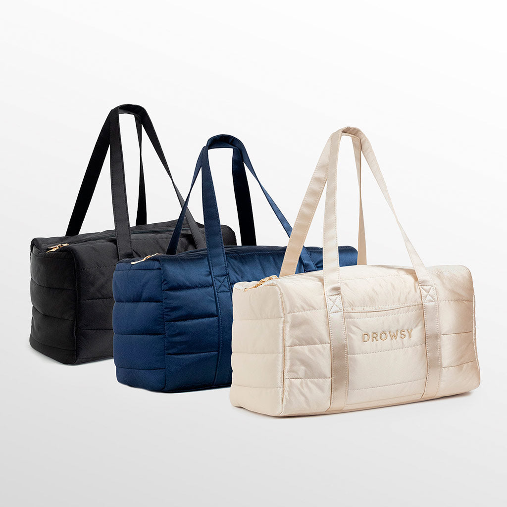 Three quilted duffel bags in black, navy blue, and beige are displayed side by side on a light background. The beige bag has DROWSY written on the front in gold letters.