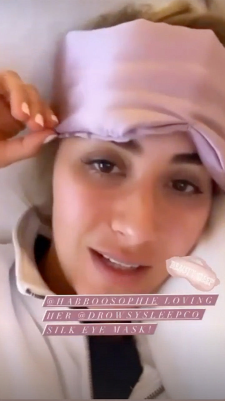 A woman lying down wears a light purple silk eye mask pushed up on her forehead. She smiles slightly and holds the edge of the mask with one hand. Text overlays mention loving a silk eye mask by Drowsy Sleep Co.