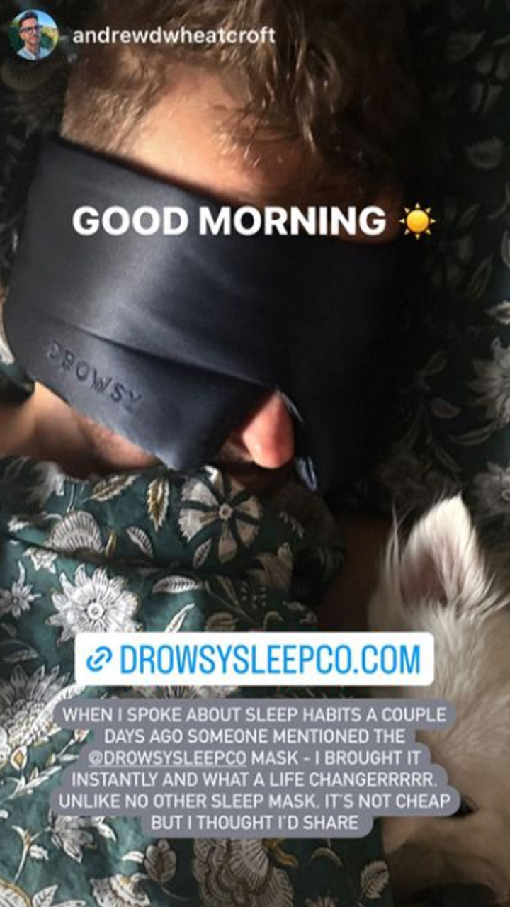A person wearing a black Drowsy sleep mask lies in bed under a patterned blanket with a small light-colored dog nearby. Overlaid text describes the mask as a life changer and shares a link to drowsysleepco.com.