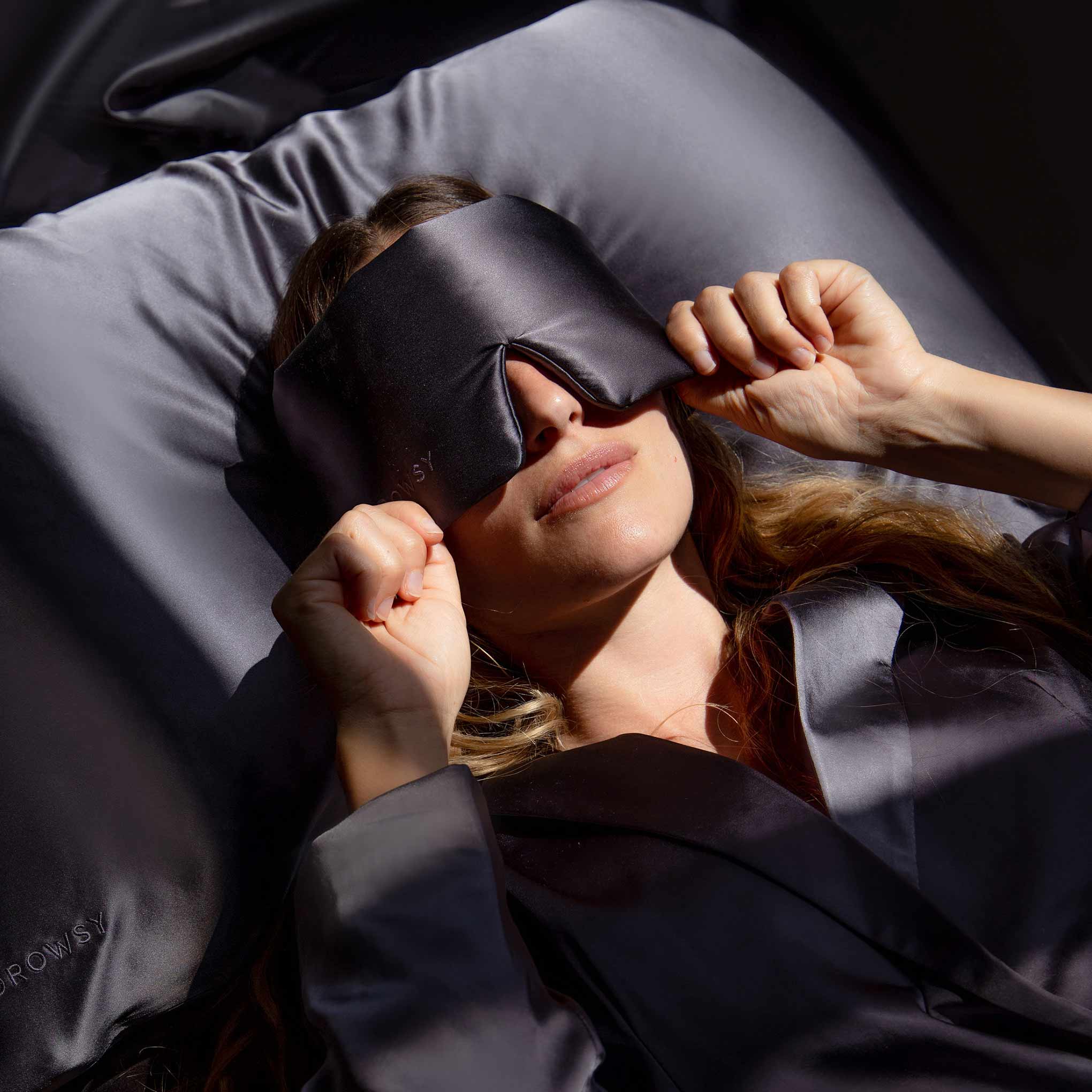 A person lying on a bed with shiny dark sheets wears a dark satin sleep mask and pajamas, gently adjusting the mask with both hands. Soft daylight falls across their face.
