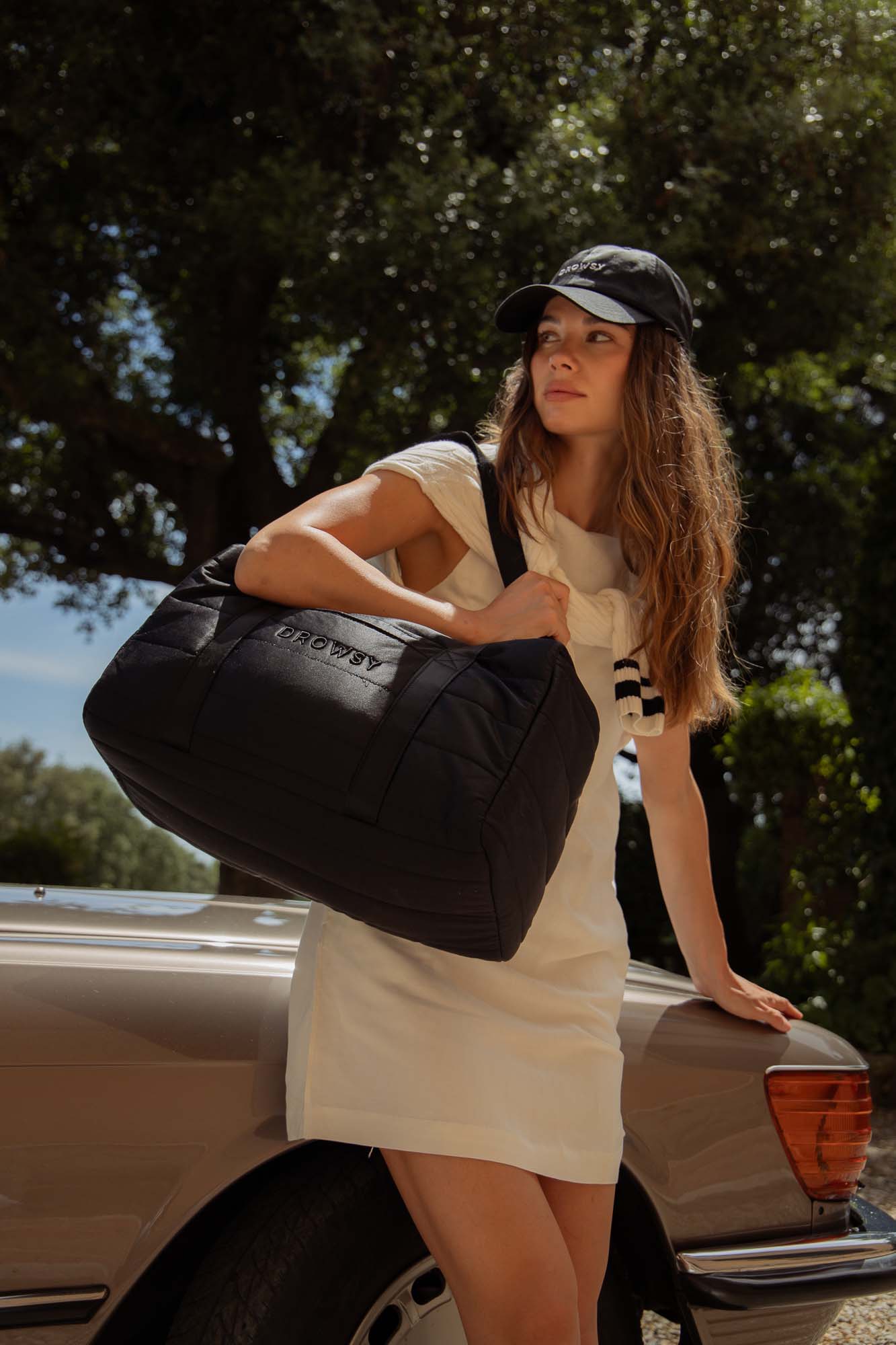 A woman in a casual white dress and black cap leans against a tan car, holding a large black duffel bag. She looks off into the distance, with trees and sunlight in the background.