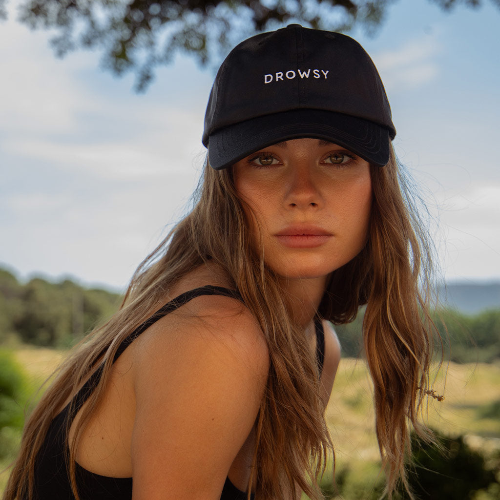 A woman with long brown hair wears the Drowsy Silk Cap - Black Jade by Drowsy and a black tank top outdoors, surrounded by trees and blue sky—ideal for outdoor workouts and preventing hat head.