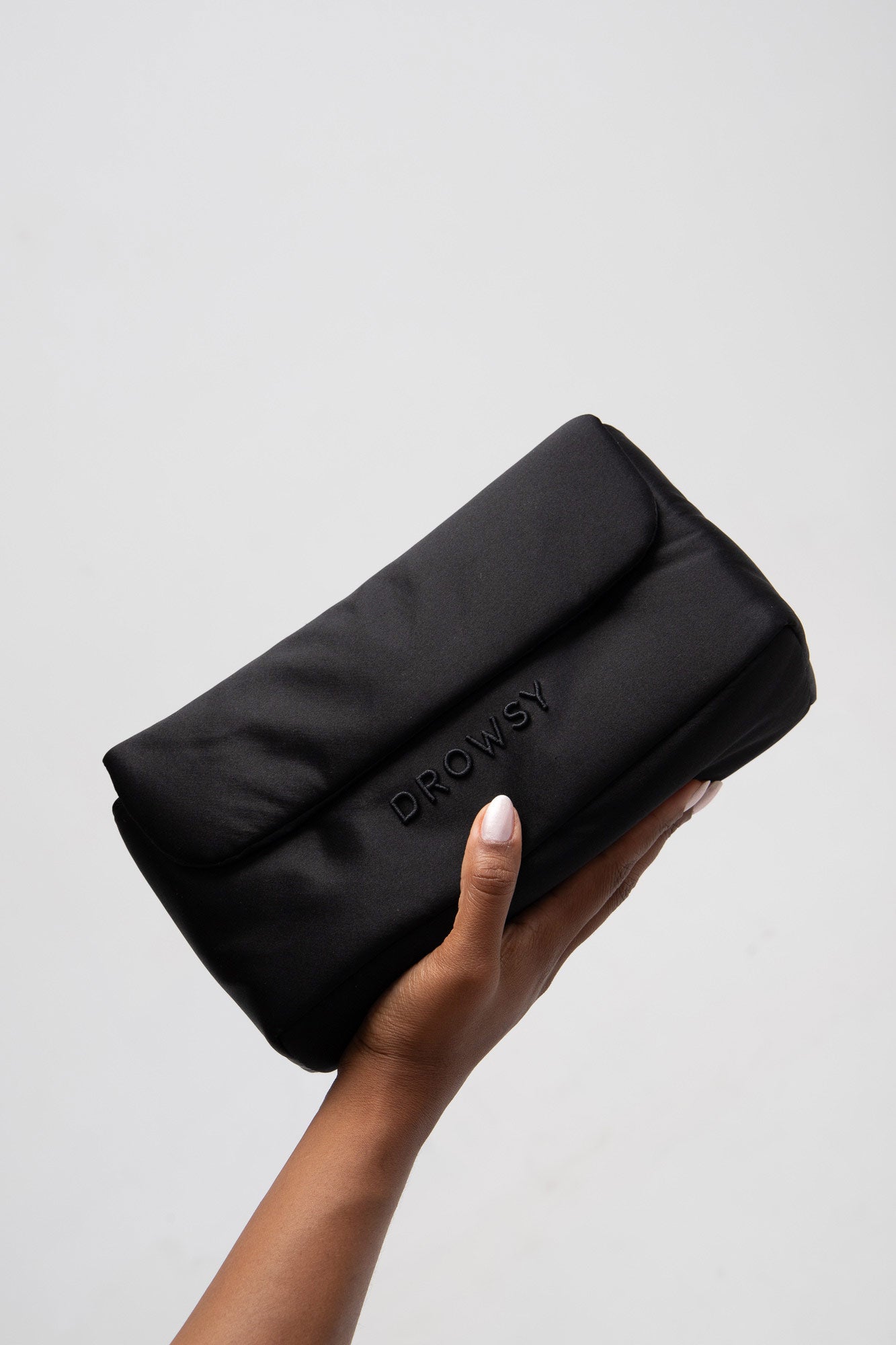 A hand holding a black, rectangular pouch with the word DROWSY embossed on the front, against a plain light background.