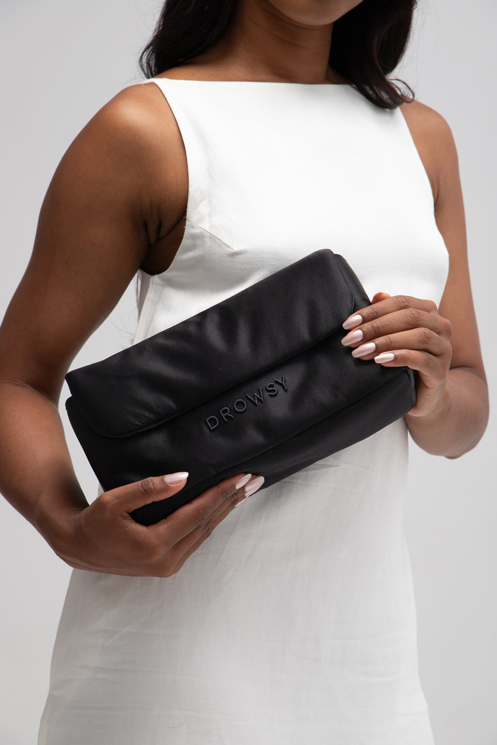 A woman in a sleeveless white dress holds a black pouch with the word DROWSY embossed on it. Only her torso and hands are visible against a plain background.