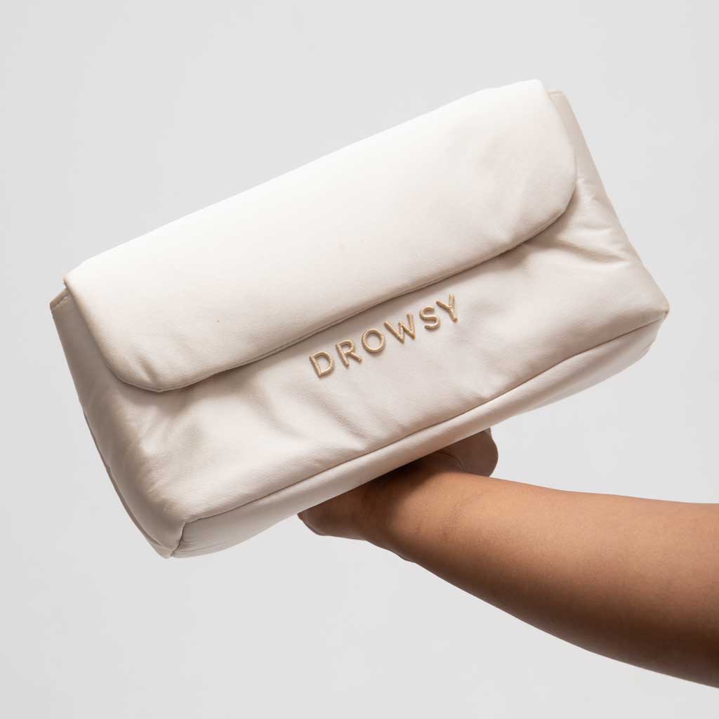 A hand holds the Drowsy Silk Necessaire in Dusty Gold by Drowsy, a soft, cream-colored pouch with gold DROWSY embroidery—an elegant accessory to elevate your beauty or skincare routine against a plain backdrop.