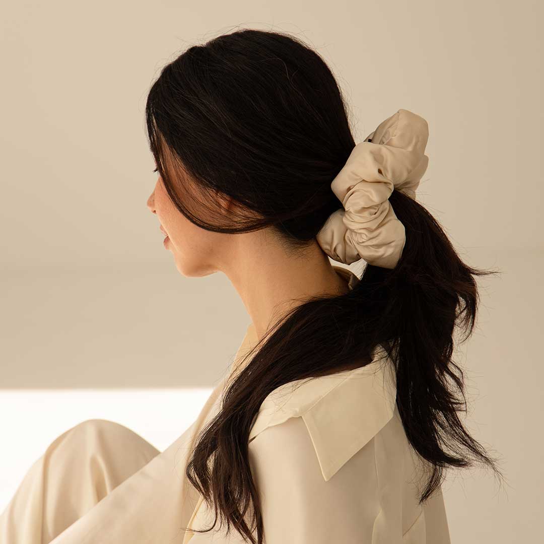 A woman with long dark hair, in a cream outfit, sits in profile with her hair tied back in a large beige scrunchie. The Sensual Sleep Bundle - Dusty Gold by us.drowsysleepco adds elegance to the softly lit, neutral background.