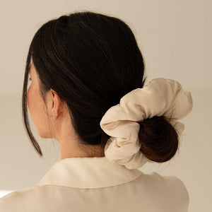 A woman with dark hair in a low bun, secured by the us.drowsysleepco Dusty Gold - Padded Scrunchie, is seen from behind wearing a cream blouse against a neutral background.