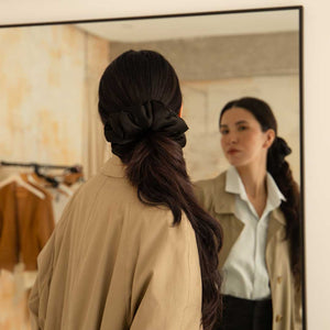 A woman with long dark hair tied in the us.drowsysleepco Black Jade - Padded Scrunchie looks at herself in a mirror, wearing a beige coat over a light blue shirt. Clothes on a rack can be seen in the background.