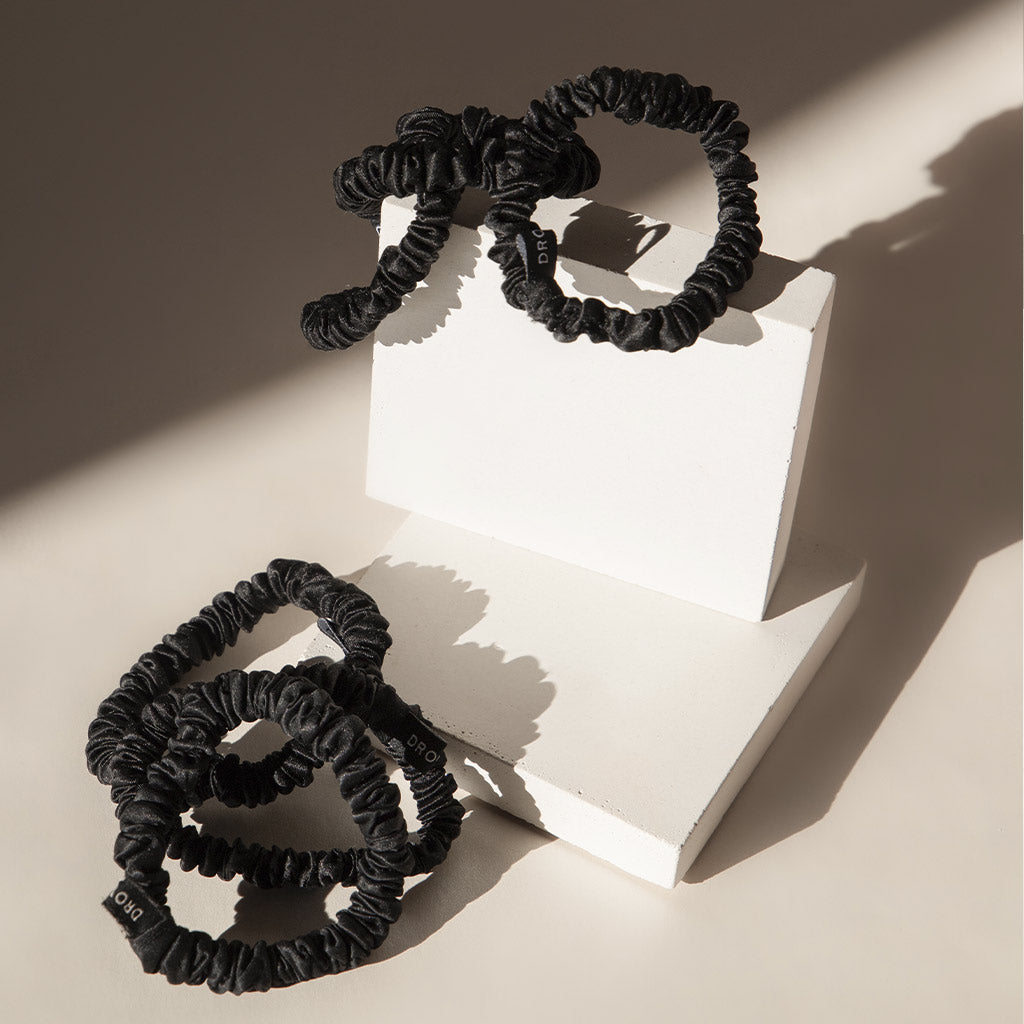 Three black scrunchies are arranged on and around two white geometric blocks, with soft shadows cast by sunlight across the minimalist scene.