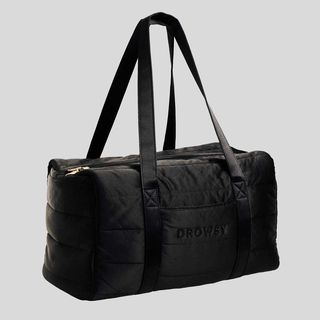 The Drowsy Silk Weekender - Black Jade by Drowsy features black quilted silk, two long shoulder straps, a gold zipper, and DROWSY embroidery on the front. This spacious bag is shown against a plain gray background.