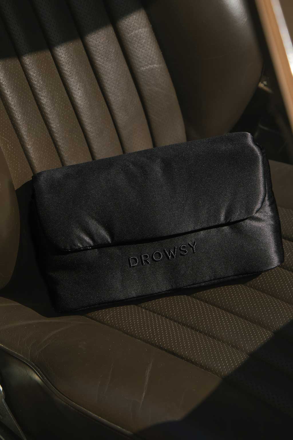 A black satin pouch with the word DROWSY embroidered on it sits on a brown leather car seat in sunlight.
