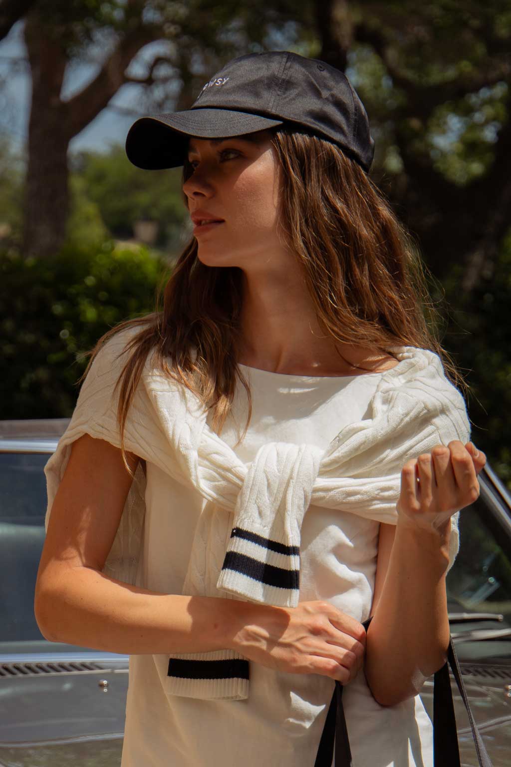 A woman wearing a black baseball cap and a white sweater draped over her shoulders stands outdoors in sunlight, holding a strap. Trees and a car are visible in the background.