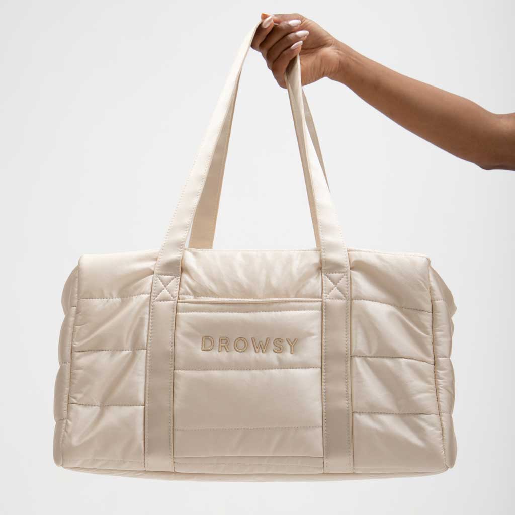 A hand holds the Drowsy Silk Weekender in Dusty Gold, a chic, cream-colored quilted duffel by Drowsy with “DROWSY” embroidered on the front pocket, featuring a spacious design against a plain light background.