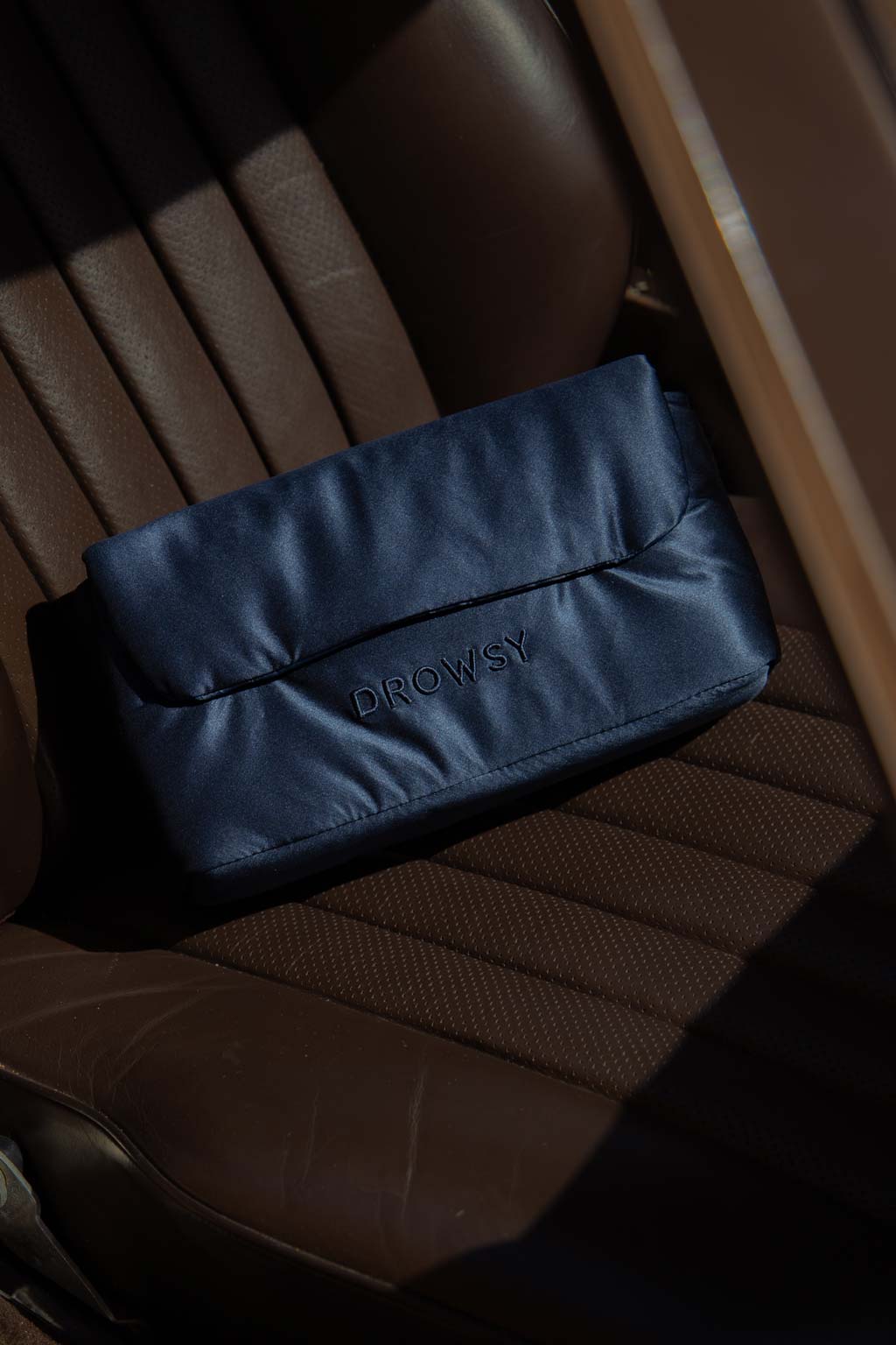 A navy blue pouch with “DROWSY” stitched on the front rests on the brown leather seat of a car, partially highlighted by sunlight.