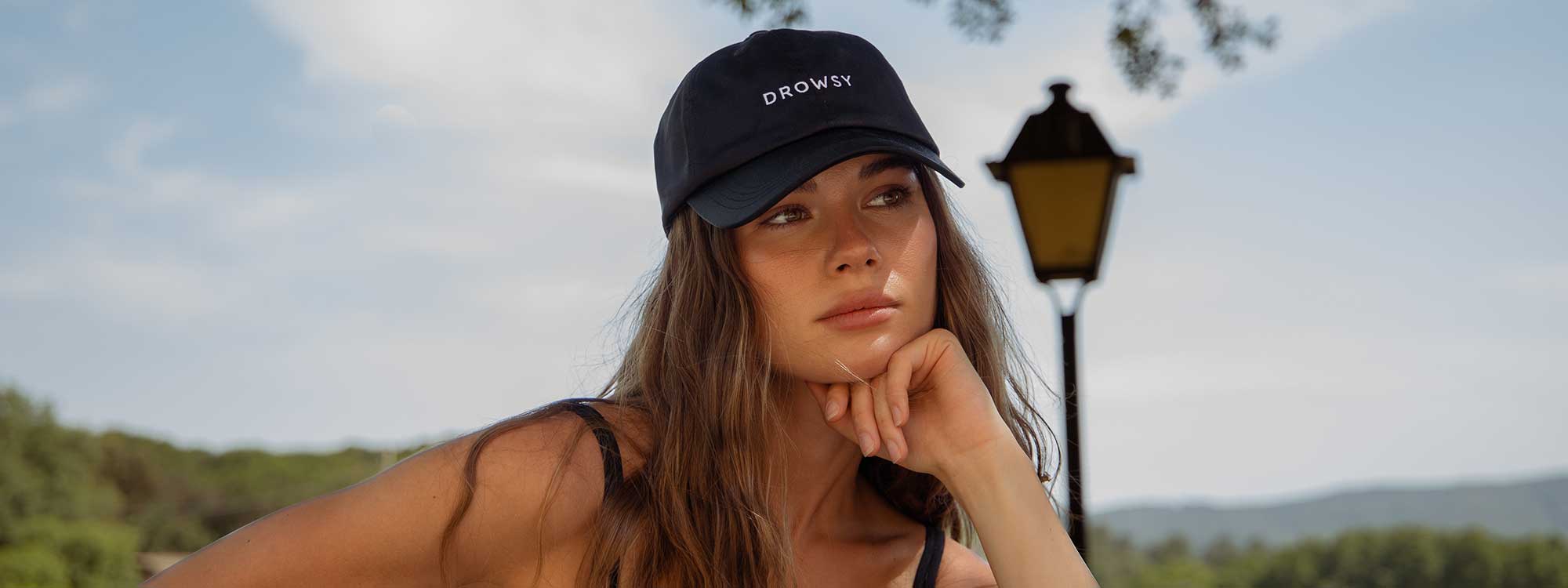 A woman with long brown hair wearing a black DROWSY cap and black tank top rests her chin on her hand outdoors, with a street lamp and trees in the background.