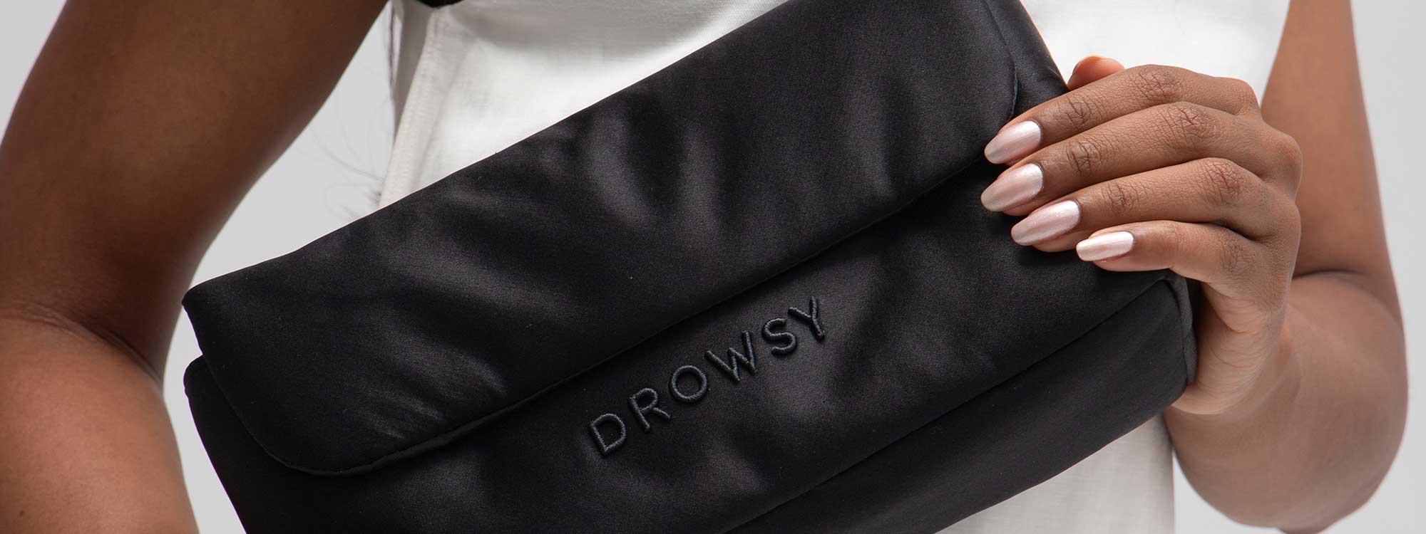 A person holds a black, soft pouch with “DROWSY” embroidered on it, against their white shirt. Their nails are manicured and painted in a light shade.