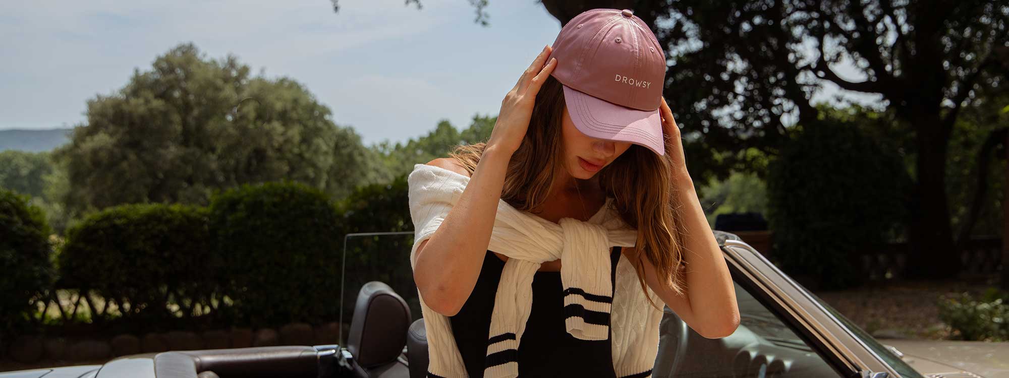 A woman stands next to a convertible car, adjusting a pink baseball cap. She wears a black top with a white sweater draped over her shoulders. Lush, green trees and bushes are visible in the background.