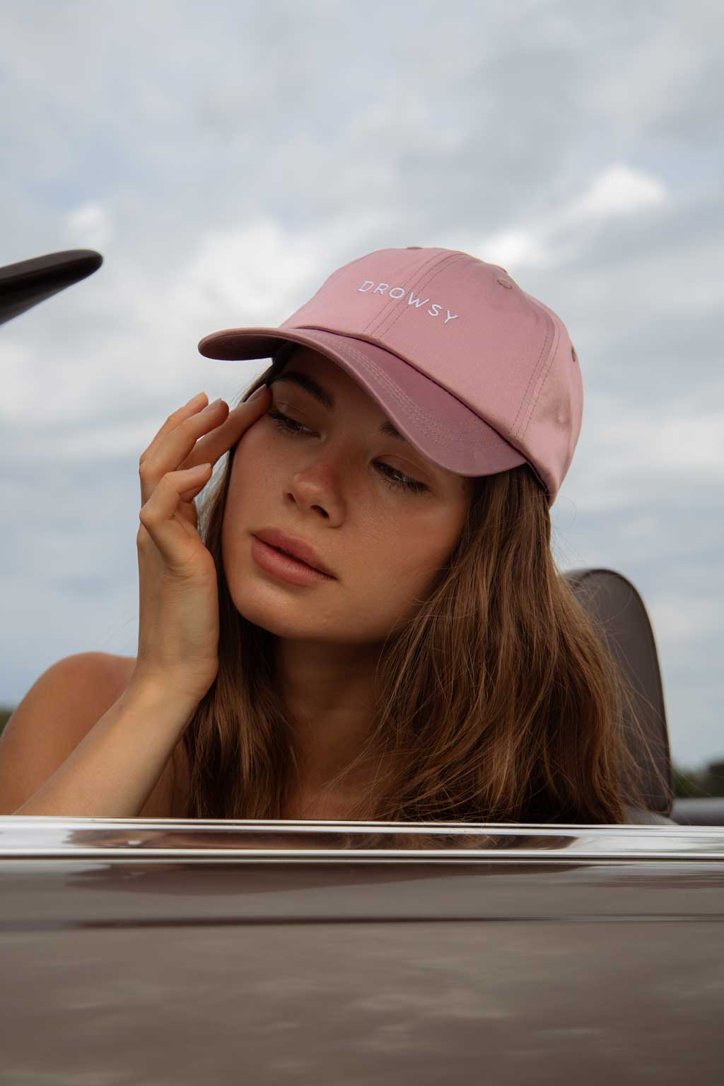 A woman with long brown hair wears a pink cap that says DROWSY. She rests her hand on her face and sits in a car with the window down, with a cloudy sky in the background.