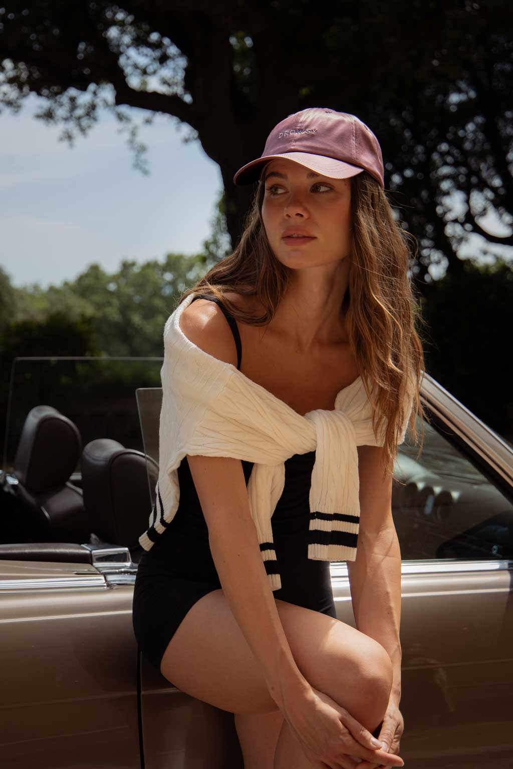 A woman in a black outfit and light sweater sits on the door of a convertible car. She wears a pink baseball cap and looks to the side, with trees and a partly cloudy sky in the background.