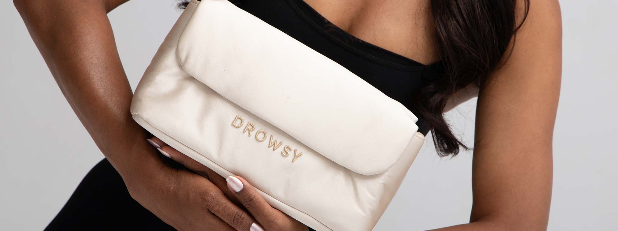 A person in a black top holds a cream-colored, rectangular pouch with the word DROWSY embroidered on it in gold letters.