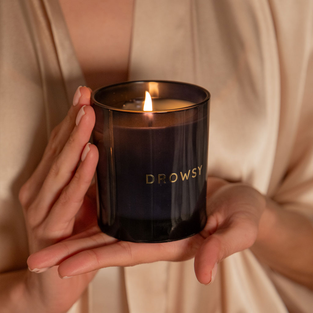 A person in a beige robe holds the drowsysleepco Sleep Ritual Candle 220g, a black glass candle with DROWSY in gold letters, ideal for aromatherapy with natural essential oils.