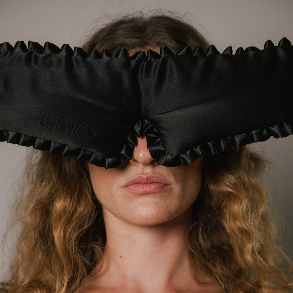 A person with wavy blonde hair wears a black, satin sleep mask with ruffled edges that covers their eyes and nose; the mask has DROWSY embroidered on it.