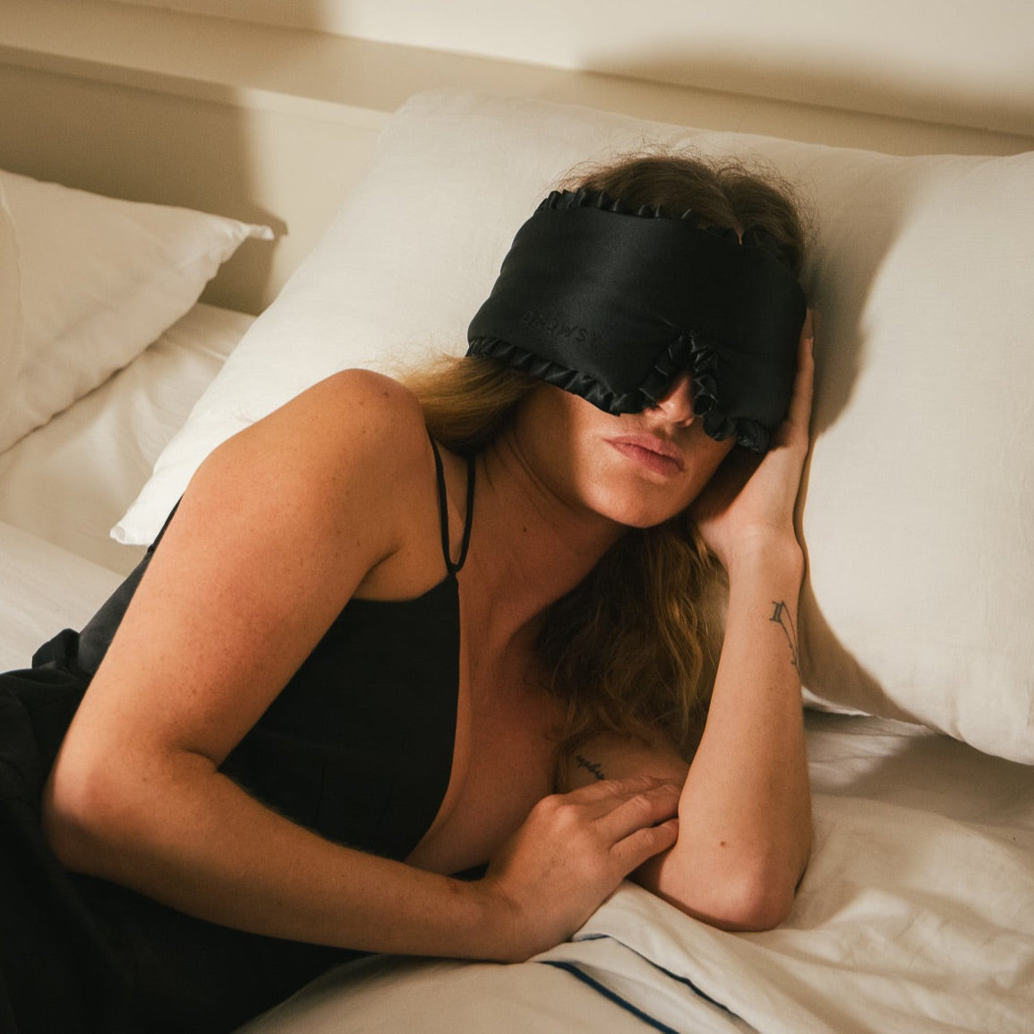 A woman with long hair lies on a bed, wearing a black sleep mask and a black camisole. She rests her head on one hand and is surrounded by white pillows and bedding.