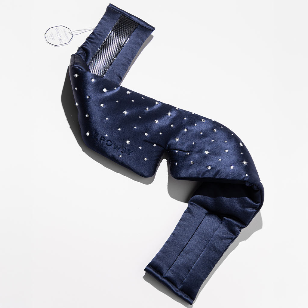 The us.drowsysleepco Silk Sleep Eye Mask Crystals by Swarovski® in navy blue with small white dots is shown unfolded on a white surface, revealing its padded sections and fabric tag. The design ensures 100% blackout for restful sleep.