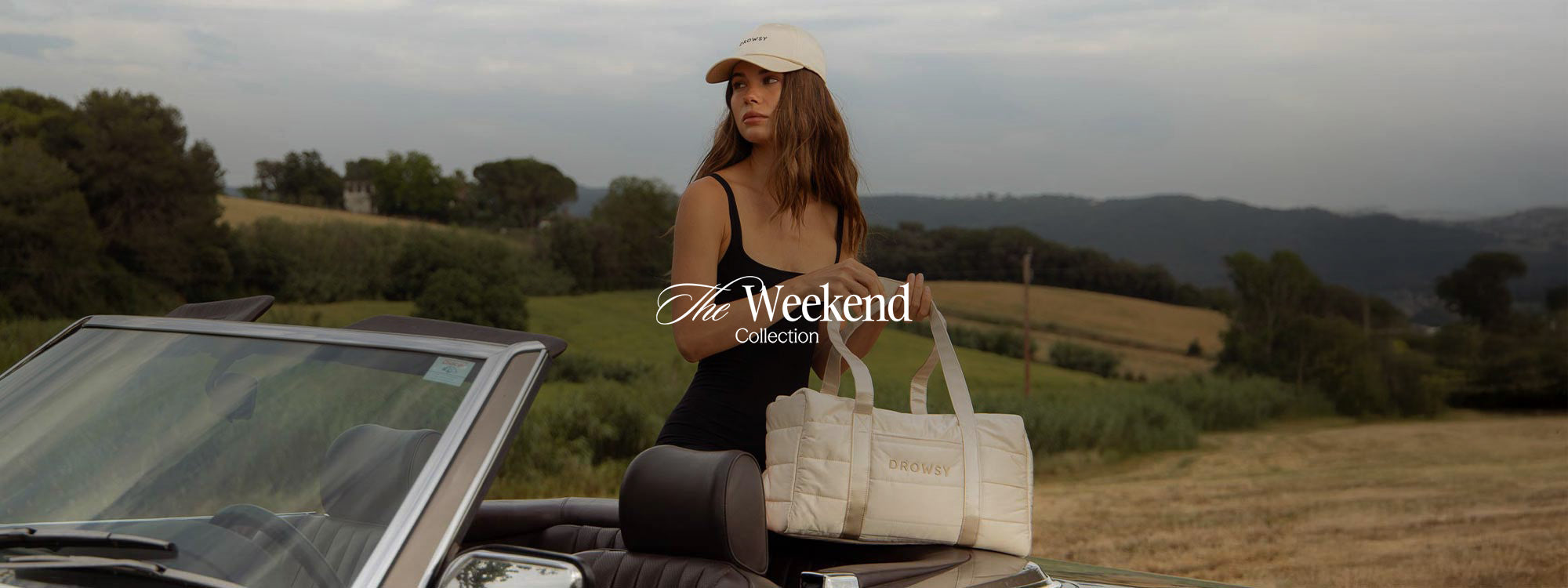 A woman in a black dress and beige cap stands beside a convertible car in a rural landscape, holding a cream-colored bag. The text The Weekend Collection is overlaid in the center.