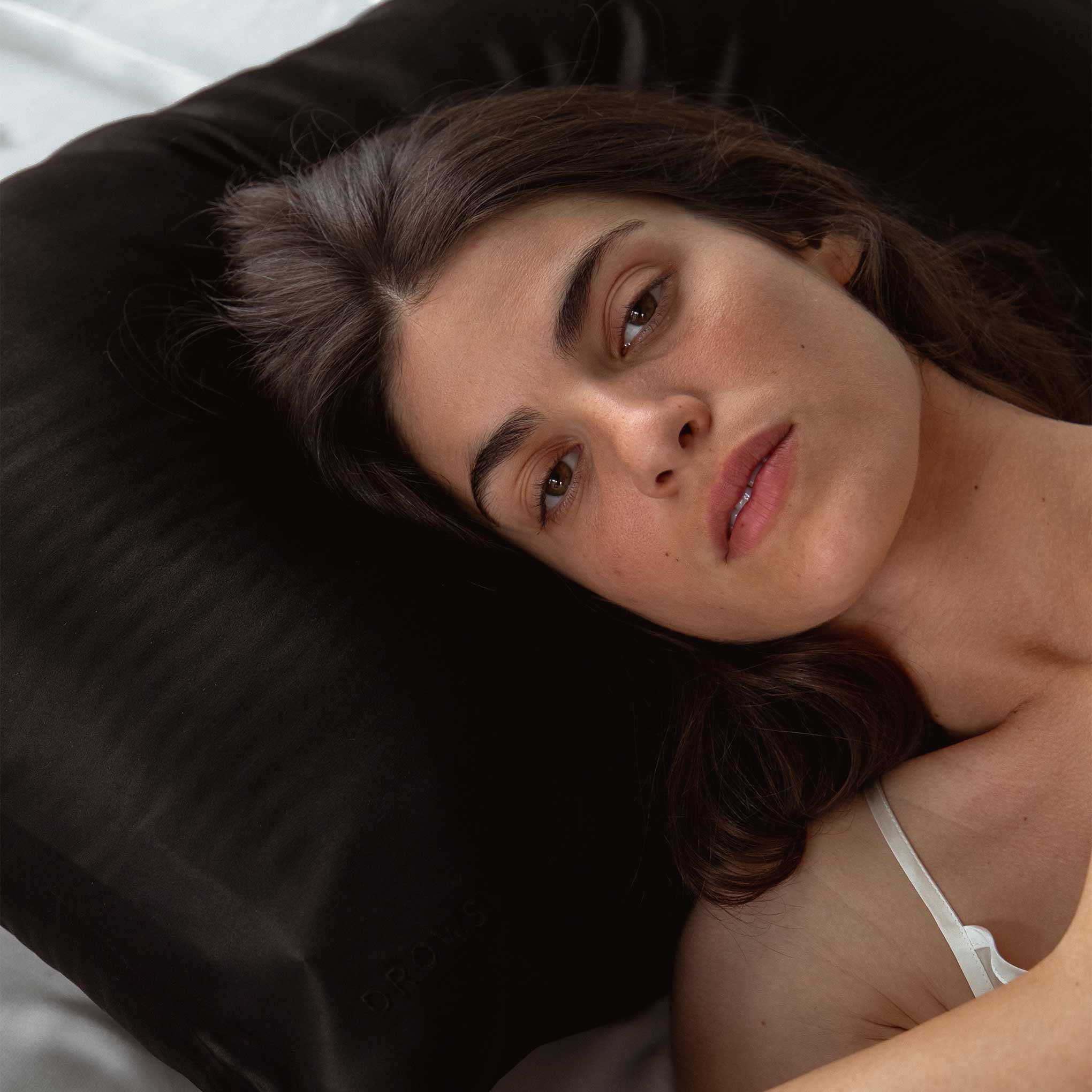 A woman with long brown hair relaxes on a drowsysleepco Sensual Sleep Bundle - Black Jade silk pillowcase, looking at the camera. She wears a white strap, while the blurred background highlights this elegant beauty sleep moment.
