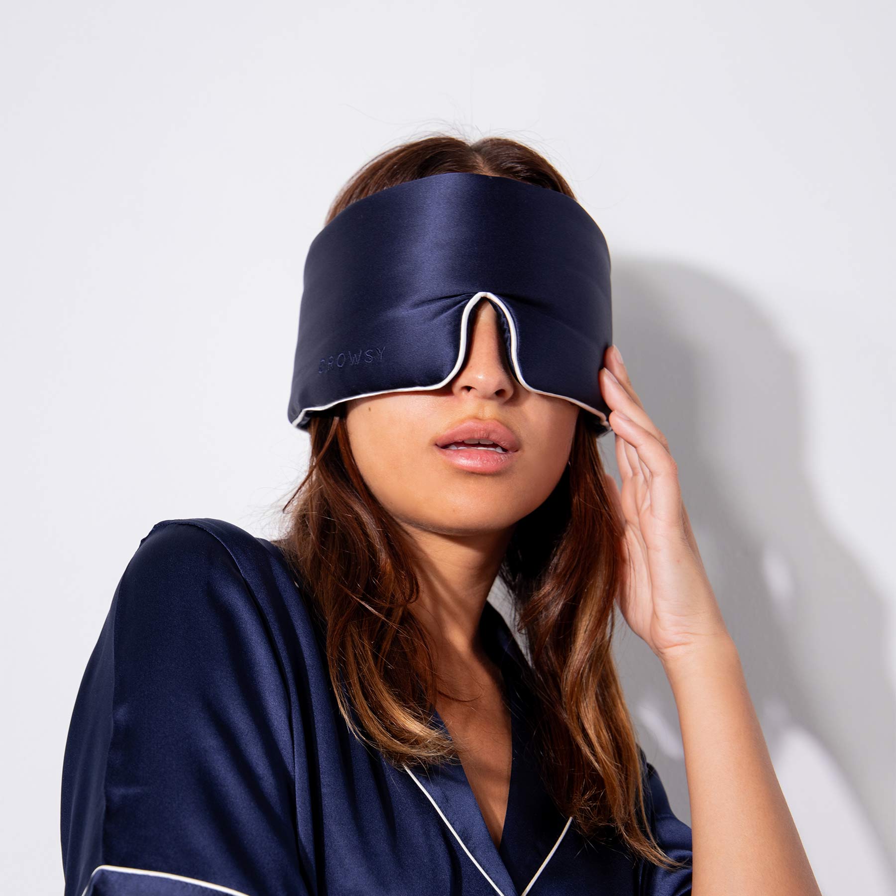 A woman in us.drowsysleepcos Midnight Blue - Pipe Dreams Edition satin pajamas and silk sleep mask stands against a white background, adjusting the mask over her eyes.