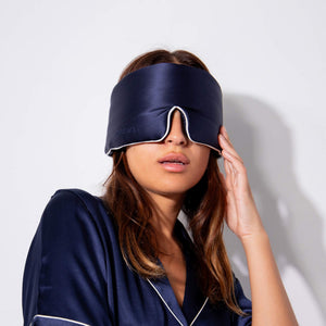 A woman in us.drowsysleepcos Midnight Blue - Pipe Dreams Edition satin pajamas and silk sleep mask stands against a white background, adjusting the mask over her eyes.