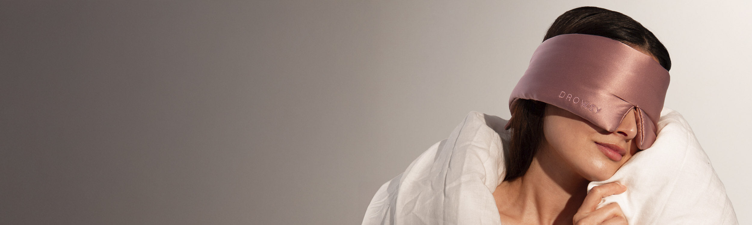 A person wearing a silky, mauve eye mask reclines with a soft white blanket wrapped around their shoulders, resting their head gently on the blanket. The background is a smooth gradient from gray to white.